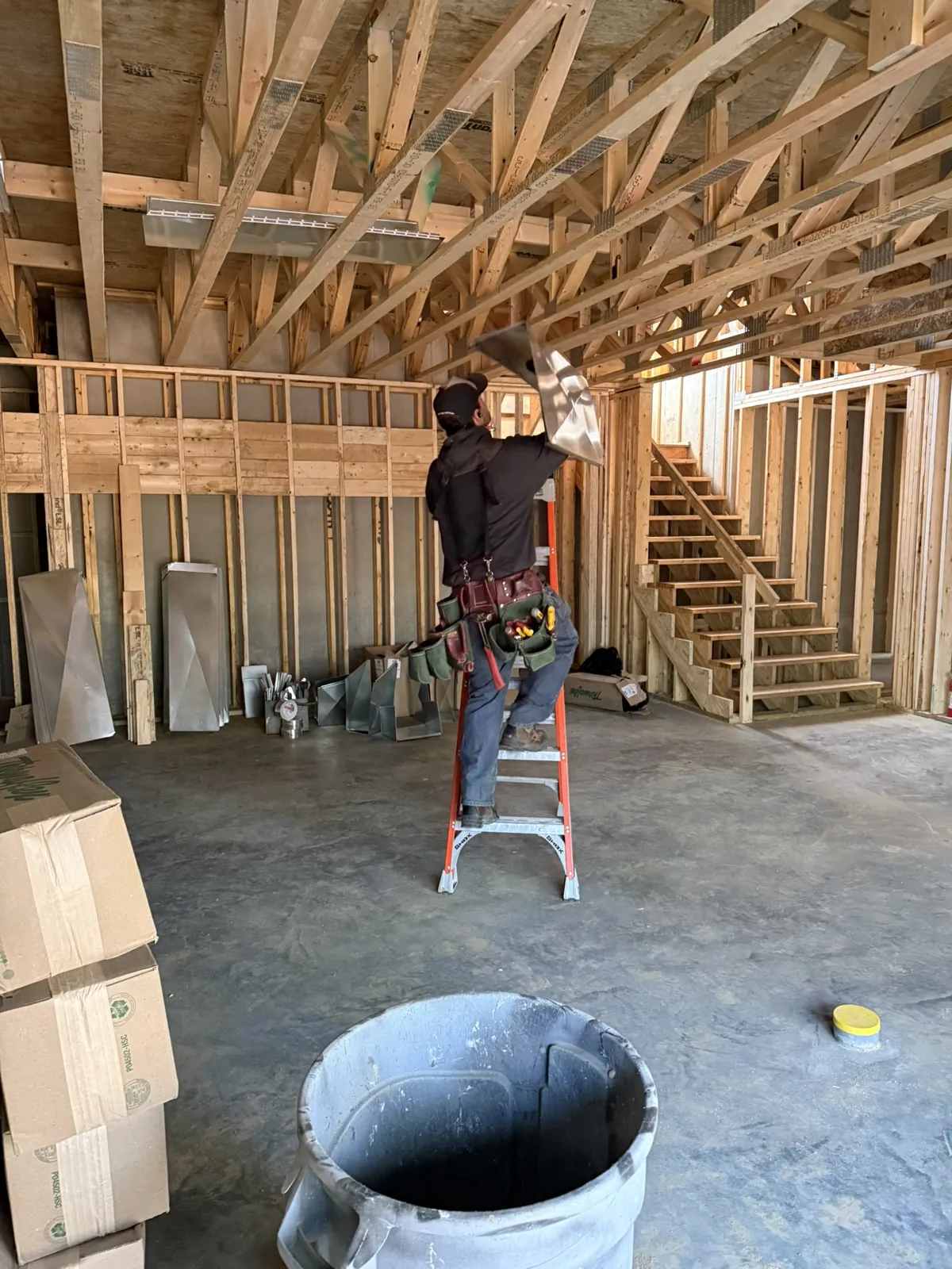 Technician on a ladder installing ductwork in a large open-framed new construction space