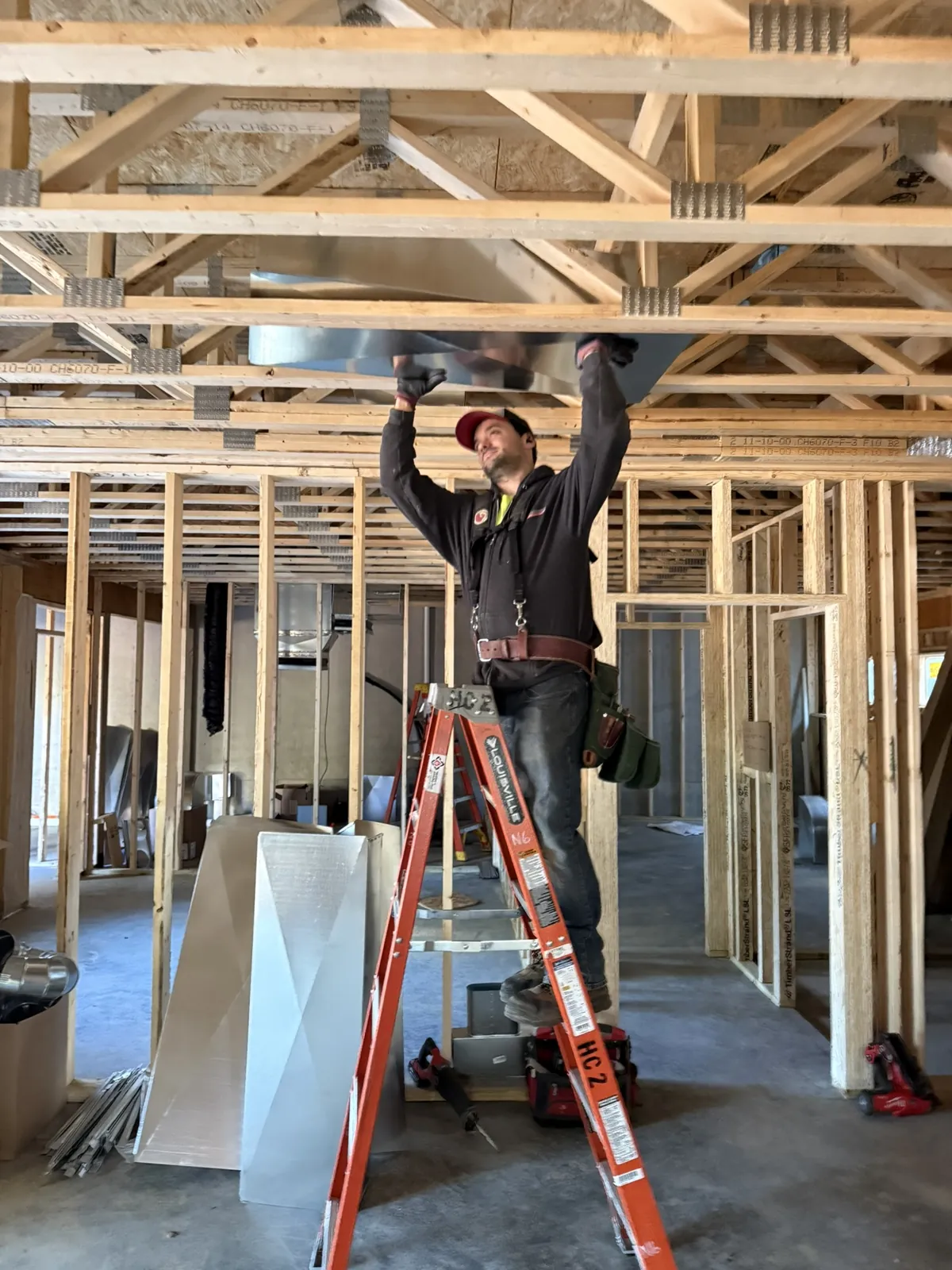 Technician on a ladder installing ductwork in the ceiling trusses of a new construction building