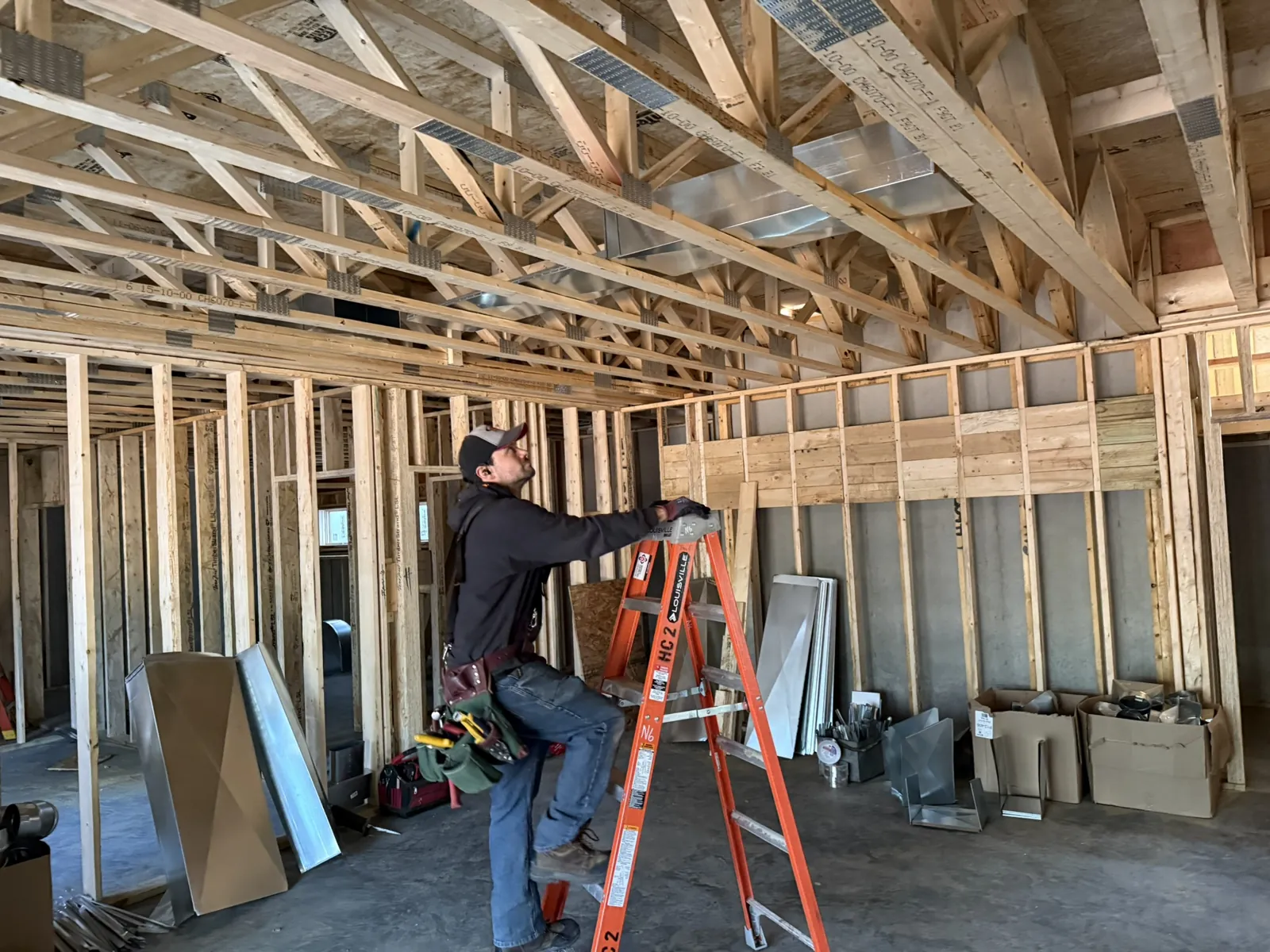 Technician on a ladder running ductwork across open ceiling trusses in a new build