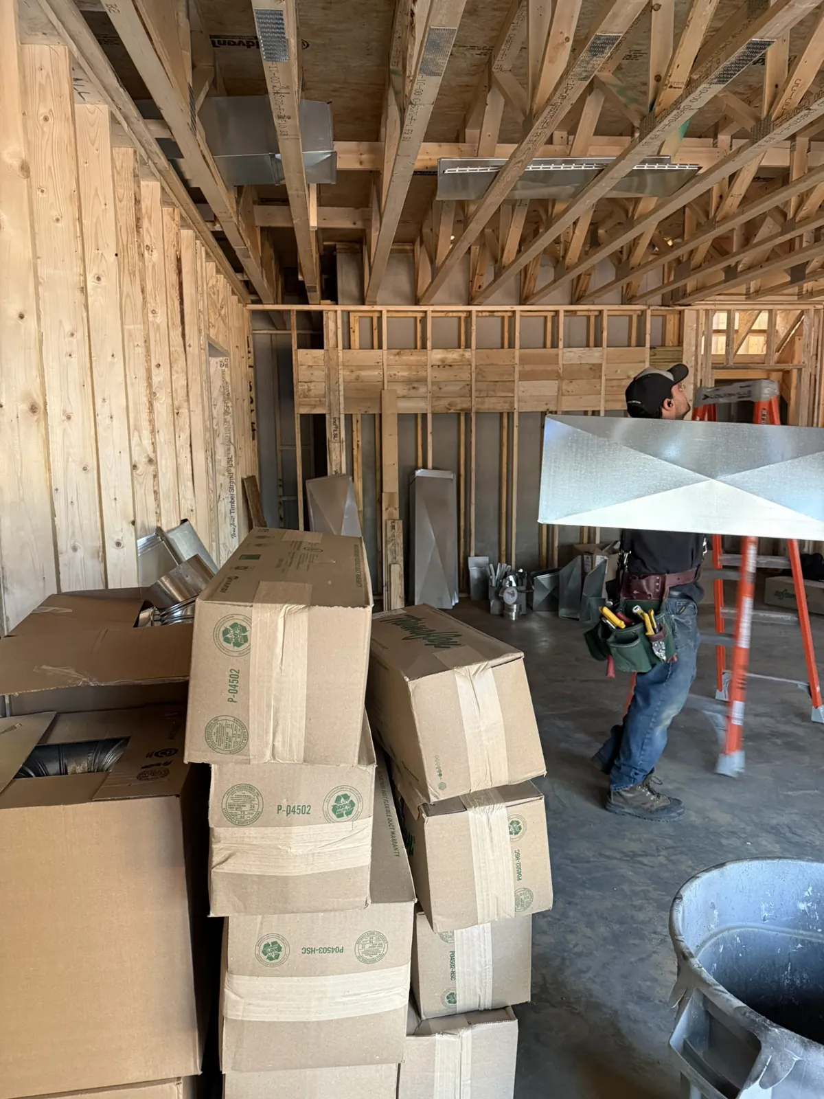 Technician carrying a sheet metal duct section past boxes of HVAC fittings on a new build